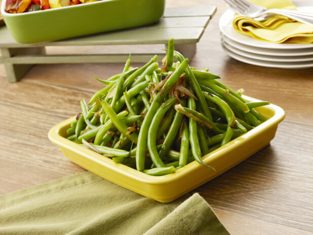 Yellow bowl of cooked green beans with caramelized onions on a wood table