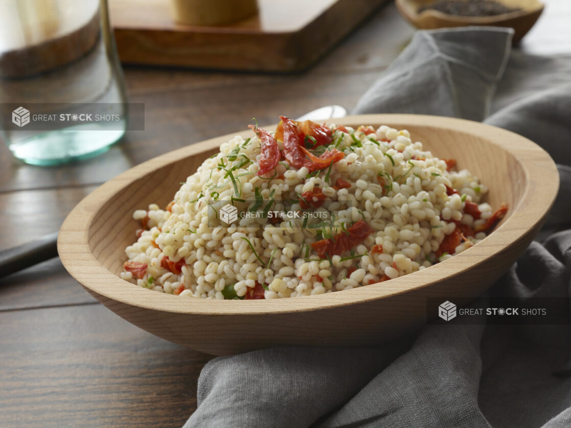 Wooden bowl of Israeli couscous salad with sundried tomatoes and fresh shredded basil tossed in a vinaigrette on a wood table with napkin