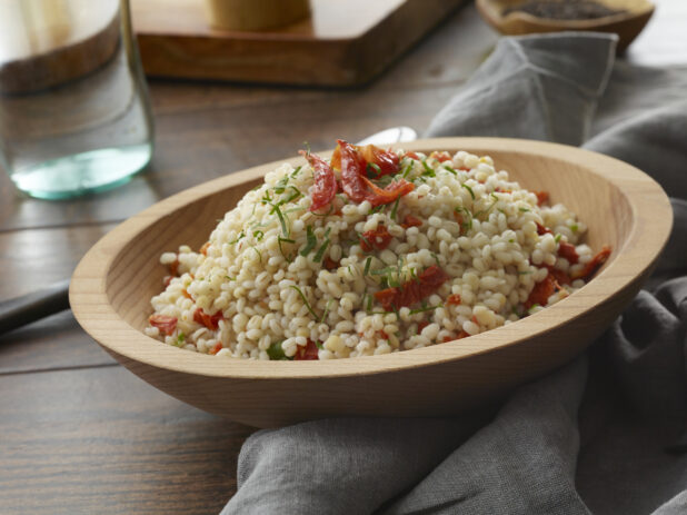 Wooden bowl of Israeli couscous salad with sundried tomatoes and fresh shredded basil tossed in a vinaigrette on a wood table with napkin