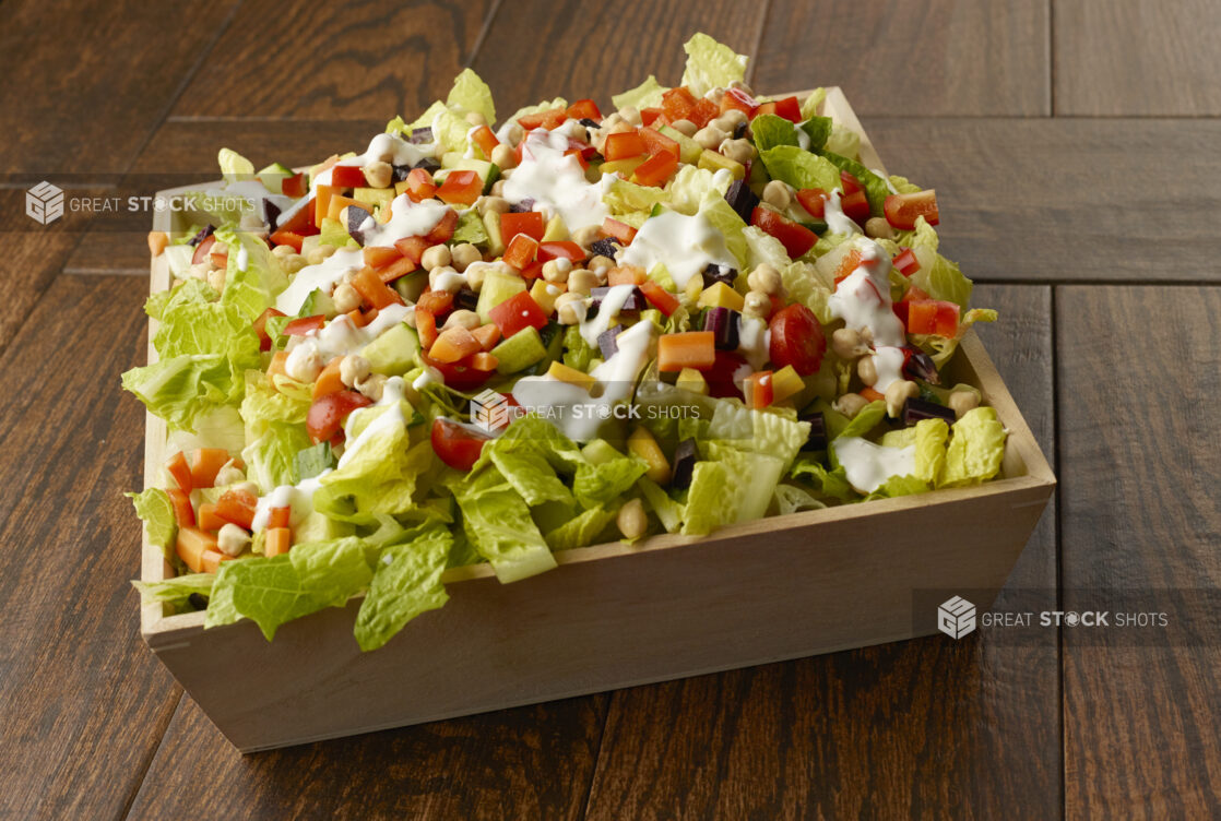 Large salad with romaine, chick peas, peppers, cherry tomatoes, beets, carrots and cucumber in a wooden catering bowl drizzled with a creamy dressing on a wooden table