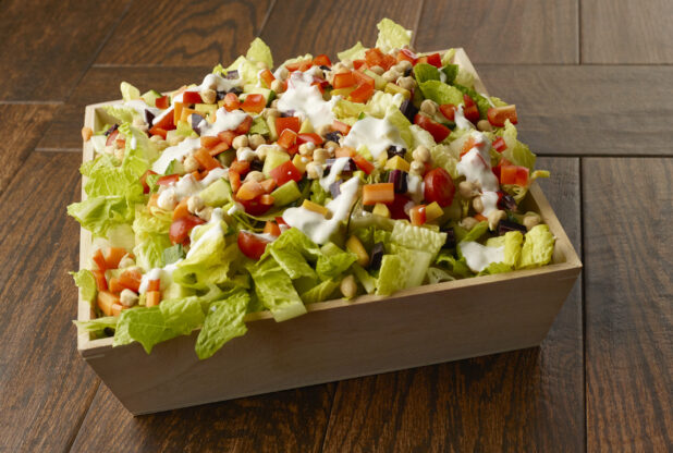 Large salad with romaine, chick peas, peppers, cherry tomatoes, beets, carrots and cucumber in a wooden catering bowl drizzled with a creamy dressing on a wooden table