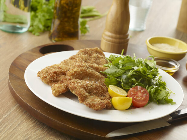 Fried schnitzel with a side arugula salad, cherry tomato and lemon on a white plate on a wooden board