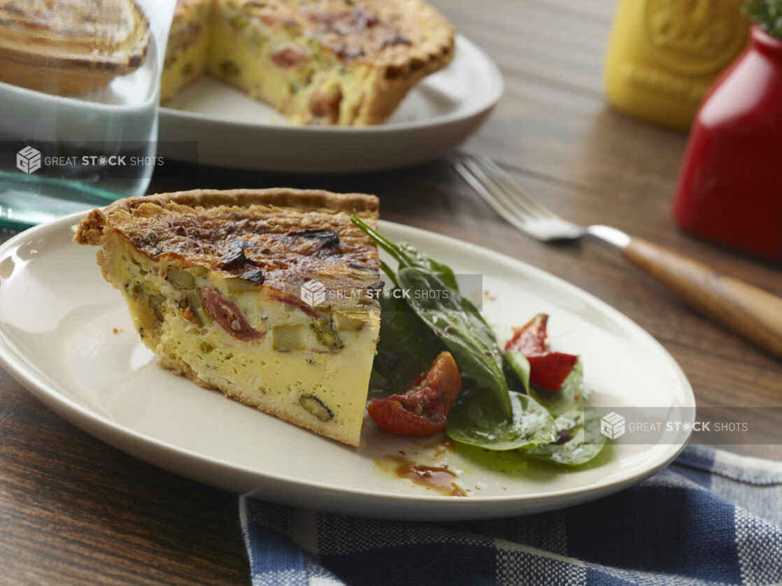 Slice of asparagus, sun-dried tomato and cheese quiche with a side salad of spinach, sun-dried tomatoes and vinaigrette with the full pie in the background on a wood table
