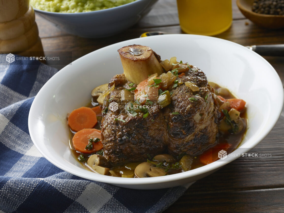 Bowl with braised veal osso buco, carrots, mushroom, celery and onions in broth on a wood table