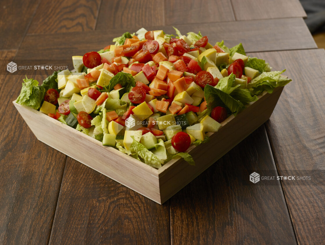 Large salad with romaine, zucchini, peppers, cherry tomatoes, carrots and cucumber in a wooden catering bowl drizzled with a vinaigrette dressing on a wooden table