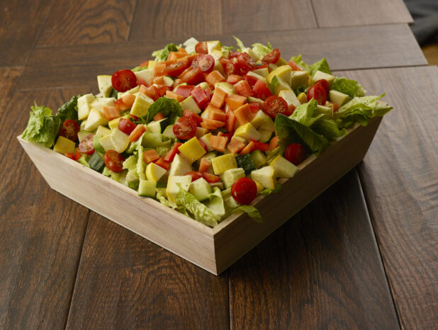 Large salad with romaine, zucchini, peppers, cherry tomatoes, carrots and cucumber in a wooden catering bowl drizzled with a vinaigrette dressing on a wooden table