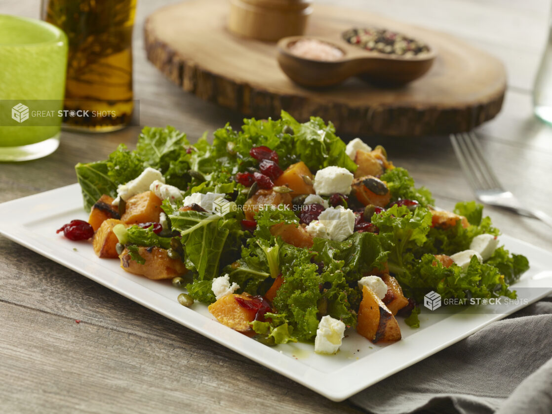 Hearty fall kale salad with goat cheese, grilled butternut squash, dried cranberries and pumpkin seeds on a white platter