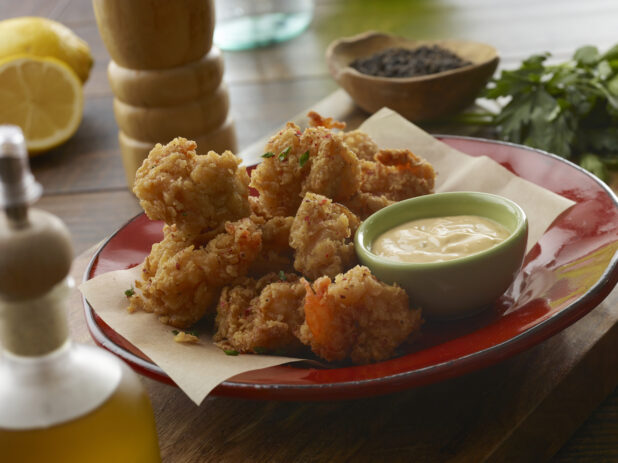 Red bowl of fried breaded popcorn shrimp with a creamy dipping sauce with fresh lemon and parsley on a wood table