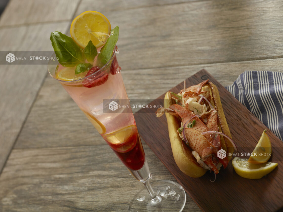 Raspberry lemonade cocktail with a lobster roll with large chunks of lobster, red onion and scallions on a toasted buttered bun with lemon wedges on a wood board