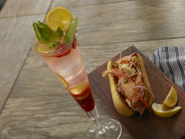 Raspberry lemonade cocktail with a lobster roll with large chunks of lobster, red onion and scallions on a toasted buttered bun with lemon wedges on a wood board