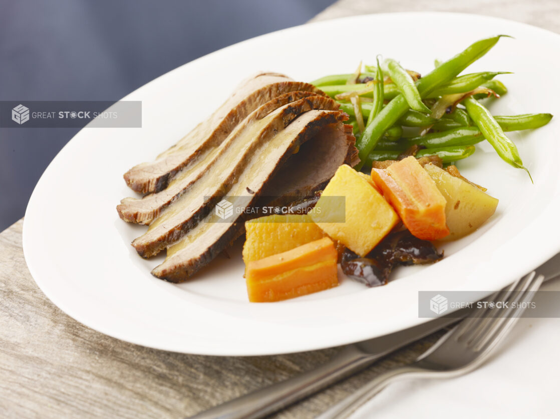 Entrée of beef brisket with gravy, green beans and root vegetables with prunes on a white plate on top of a wood table