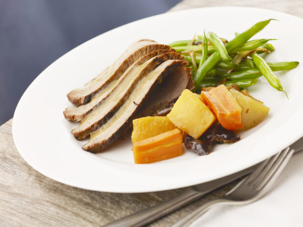 Entrée of beef brisket with gravy, green beans and root vegetables with prunes on a white plate on top of a wood table