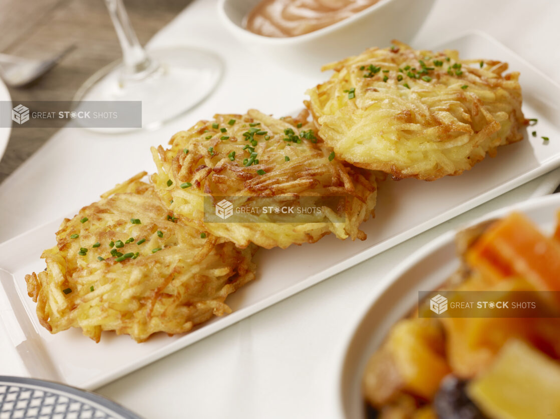 Three fried potato latkes on a white plate topped with fresh chiveson a white table cloth