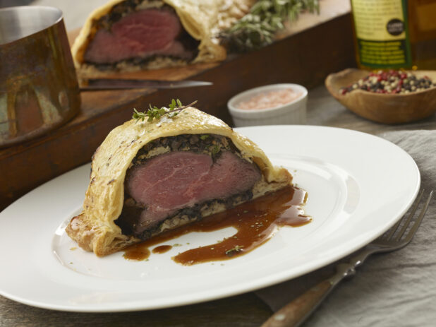 Rare beef wellington cut in half with a brown sauce on a white plate with the other half in the background