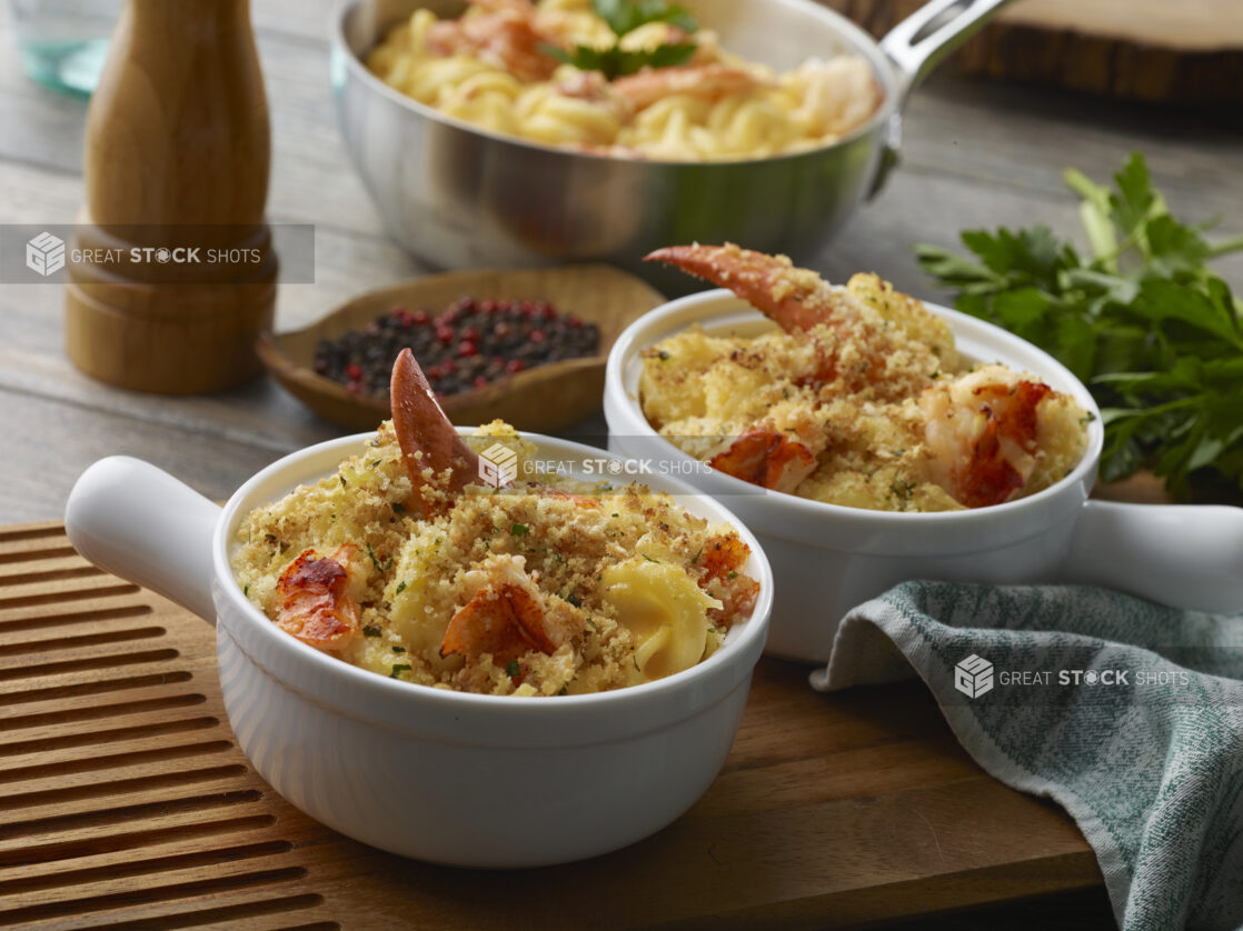 Two individually sized dishes with baked lobster mac and cheese with crumble topping on a wood board with a fresh pot of lobster mac and cheese in the background