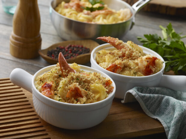 Two individually sized dishes with baked lobster mac and cheese with crumble topping on a wood board with a fresh pot of lobster mac and cheese in the background