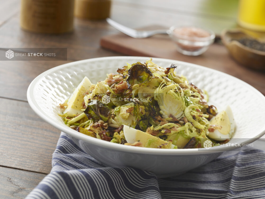 Bowl of roasted and sliced brussels sprout salad with bacon, hard-boiled eggs and sunflower seeds with a vinaigrette dressing on a wood table