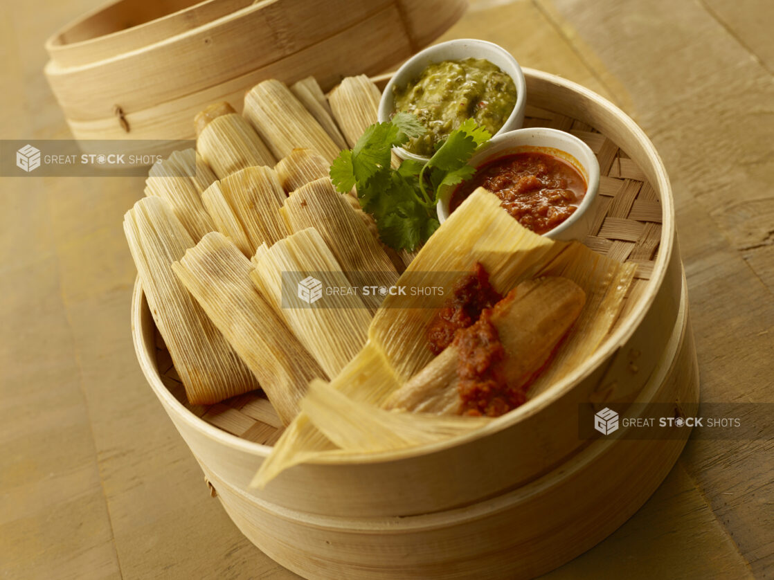 Tamales in a bamboo steamer basket with cilantro garnish and ramekins of red and green salsa on a wood tabletop