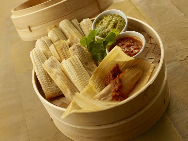 Tamales in a bamboo steamer basket with cilantro garnish and ramekins of red and green salsa on a wood tabletop