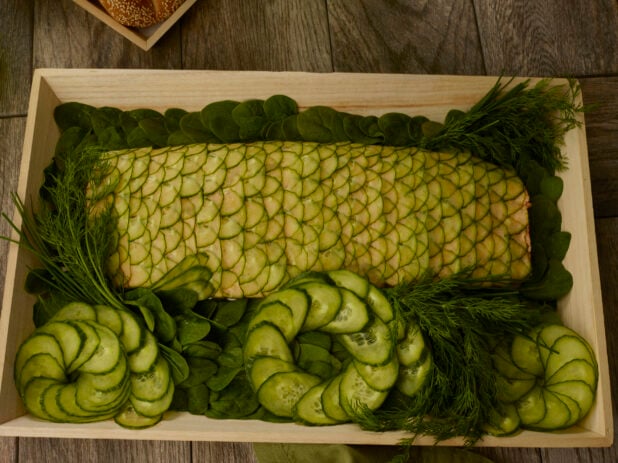 Catering presentation of cold poached salmon covered in cucumber scales on a wood catering tray with cucumber circles, spinach and fresh dill
