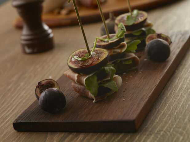 Layered appetizer of fig, prosciutto and arugula pined with a bamboo toothpick on a wooden board