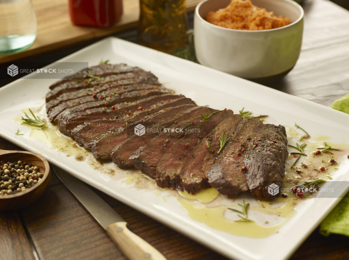 Platter of sliced medium rare flank steak on a large white platter with a bowl of pureed sweet potato in the background