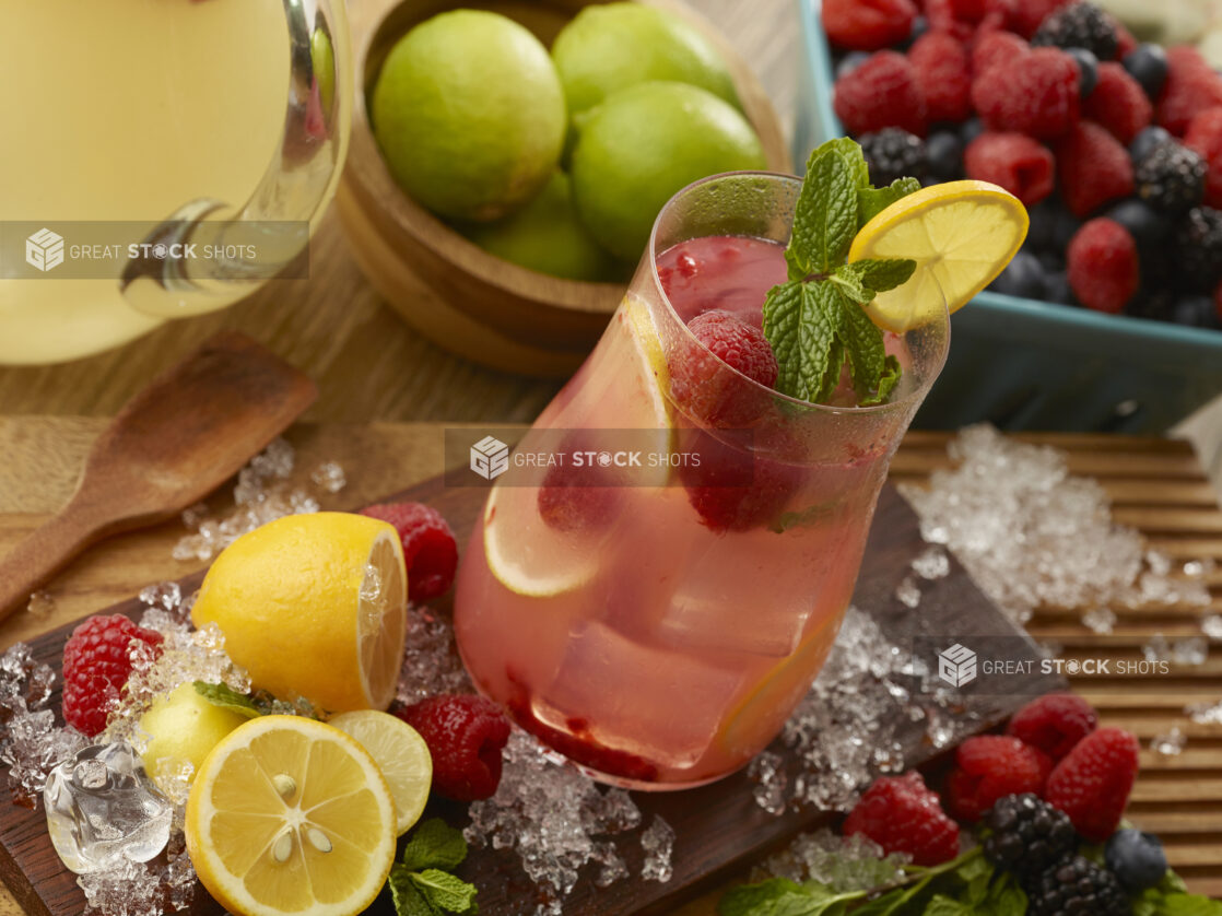 Glass of raspberry lemonade with fresh mint and a lemon wheel surrounded by crushed ice and fresh fruit on a wooden board