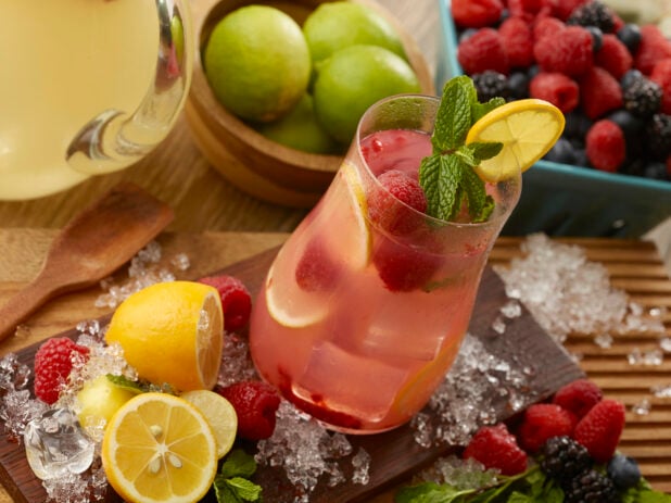 Glass of raspberry lemonade with fresh mint and a lemon wheel surrounded by crushed ice and fresh fruit on a wooden board