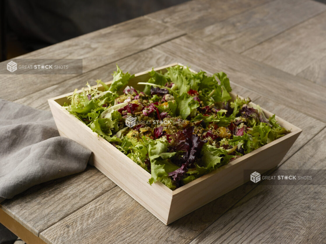 Large salad with spring mix, quinoa and roasted root vegetables in a wooden catering bowl drizzled with a vinaigrette dressing on a wooden table