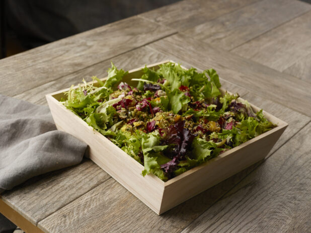 Large salad with spring mix, quinoa and roasted root vegetables in a wooden catering bowl drizzled with a vinaigrette dressing on a wooden table