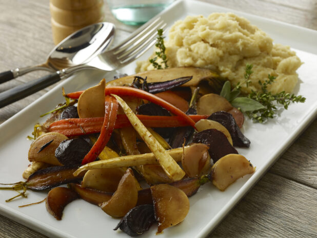 White platter of roasted root vegetables and mashed potatoes on a wood table