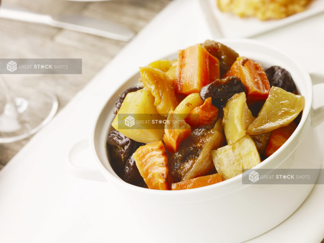 White bowl of tzimmes, potato, carrots, apples and prunes on a white table cloth