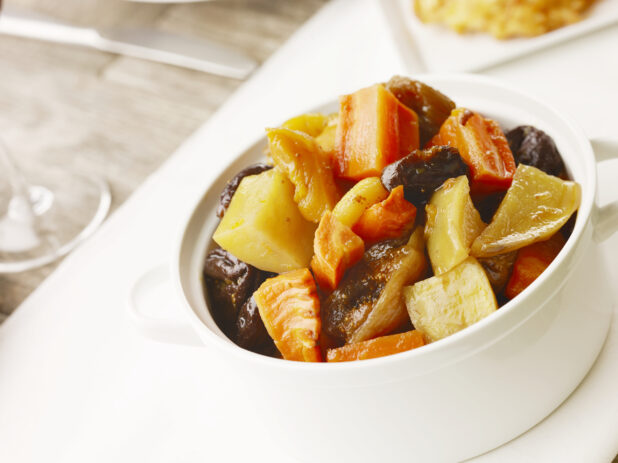 White bowl of tzimmes, potato, carrots, apples and prunes on a white table cloth