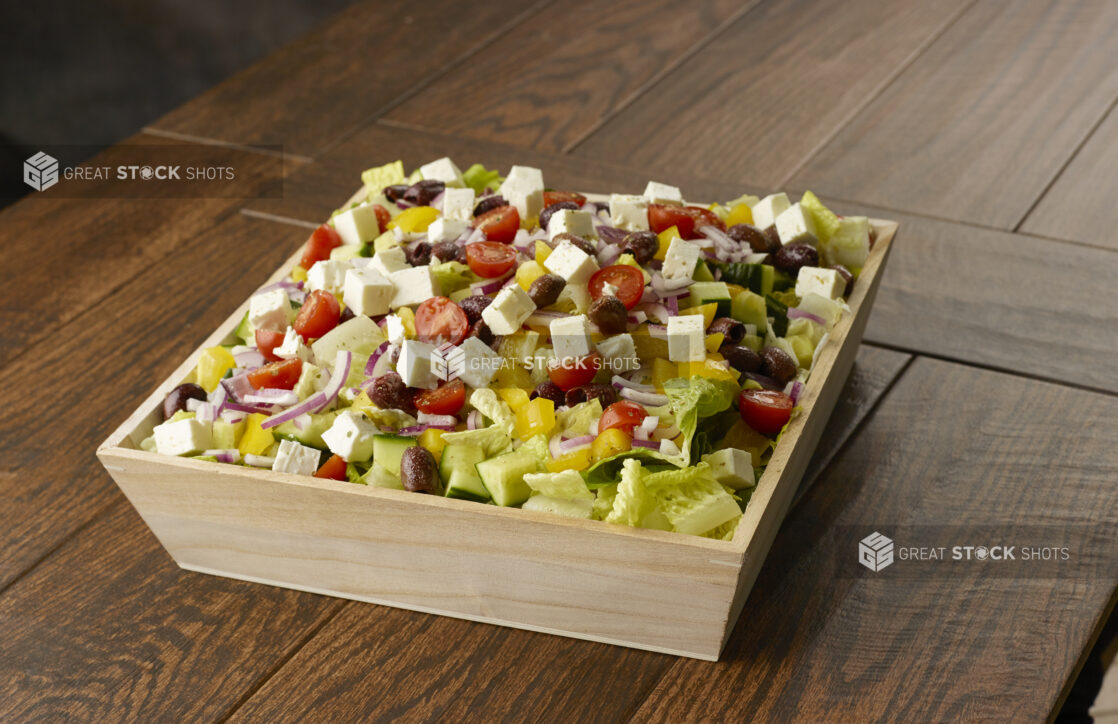 Large Greek salad with romaine, cherry tomatoes, peppers, black olives, feta, red onion and cucumber in a wooden catering bowl drizzled with a vinaigrette dressing on a wooden table