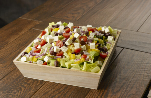 Large Greek salad with romaine, cherry tomatoes, peppers, black olives, feta, red onion and cucumber in a wooden catering bowl drizzled with a vinaigrette dressing on a wooden table