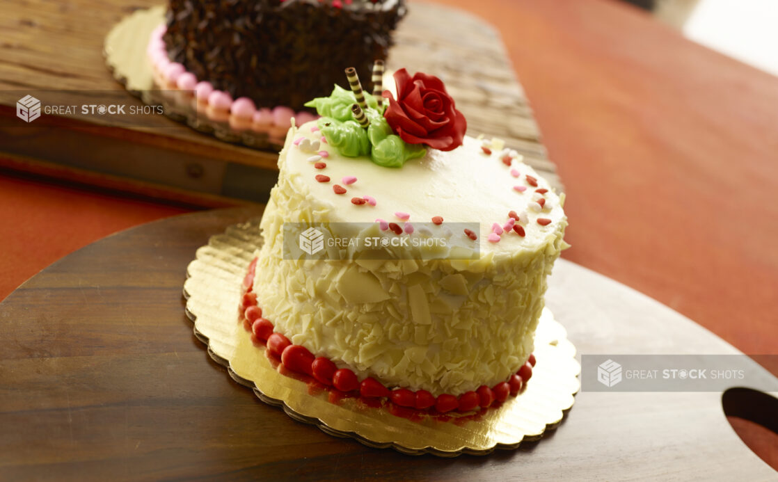 White chocolate Valentine's Day cake with heart candy and an icing sugar rose on a wooden board with a chocolate cake in the background