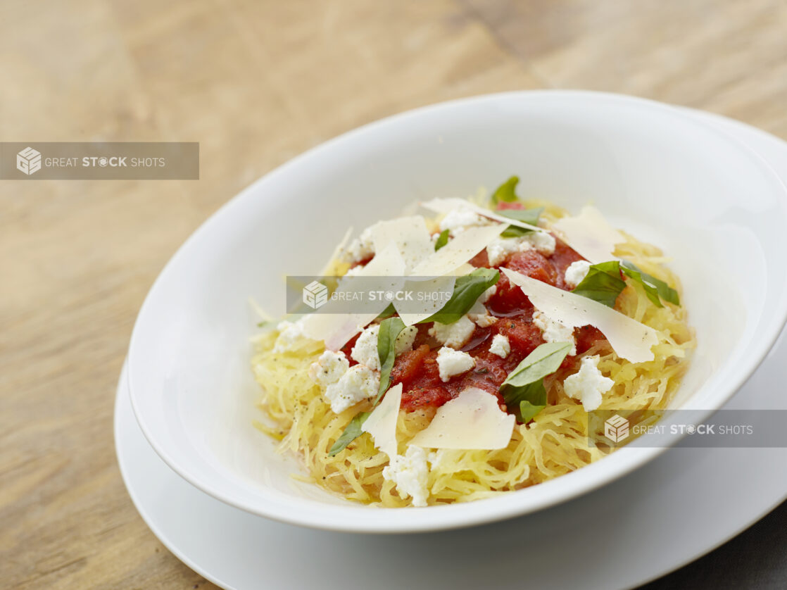 Bowl of spaghetti squash topped with tomato sauce, ricotta cheese, freshly shaved parmesan cheese and fresh basil on a wood table