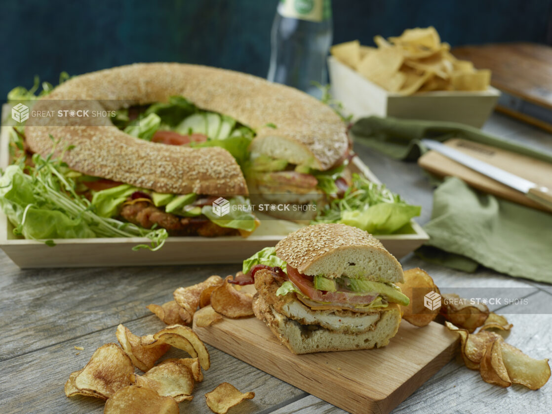 Large fried chicken sandwich ring with tomato, avocado, lettuce and bacon with a piece removed on a wooden board surrounded in kettle chips