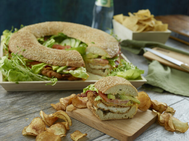 Large fried chicken sandwich ring with tomato, avocado, lettuce and bacon with a piece removed on a wooden board surrounded in kettle chips