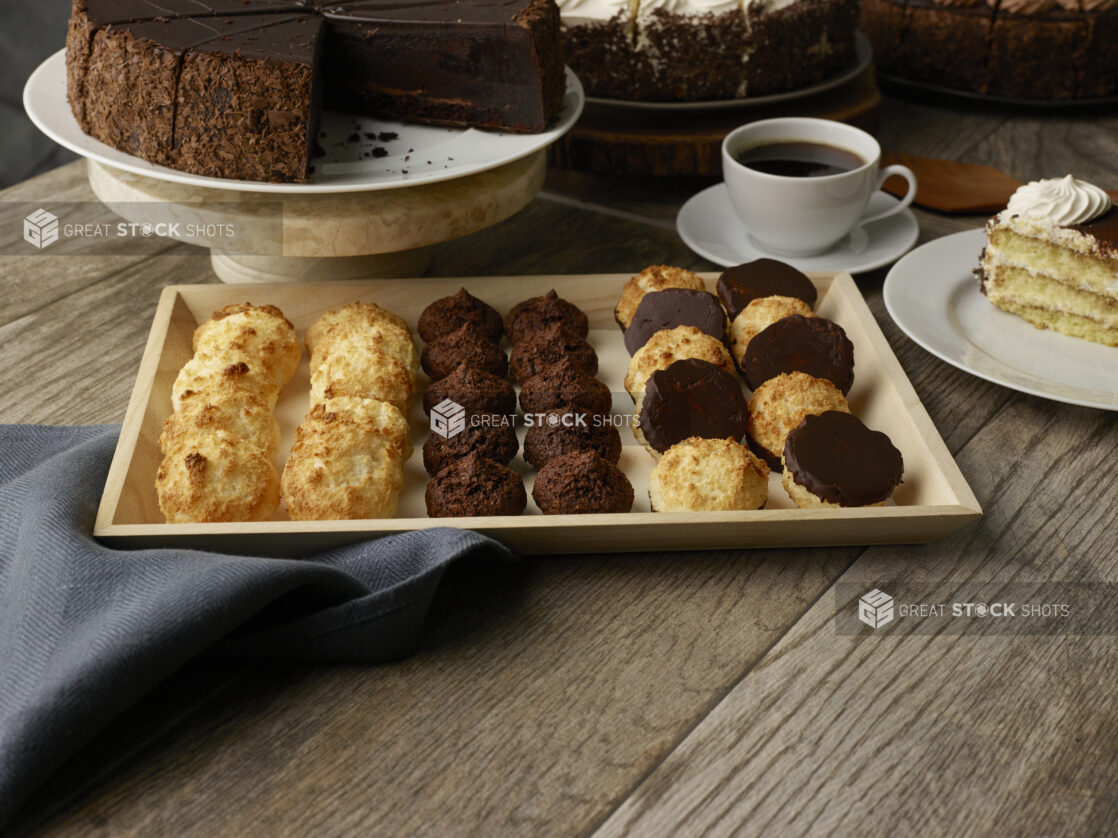 Various types of macaroons on a wood catering tray with sliced cakes in the background on stands on a wood table