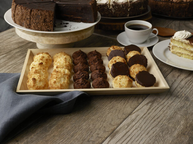 Various types of macaroons on a wood catering tray with sliced cakes in the background on stands on a wood table