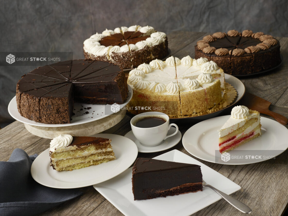 Assortment of cakes on stands and cake slices on plates with a napkin on a wood table