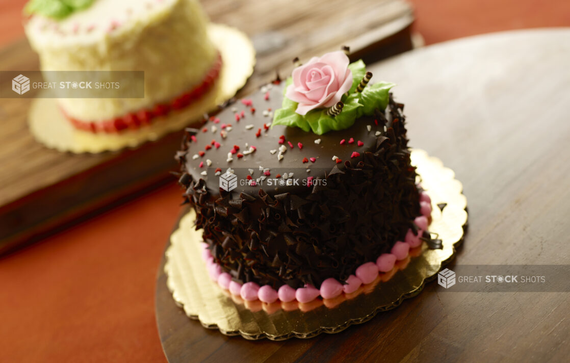 Dark chocolate Valentine’s Day cake with heart candy and an icing sugar rose on a wooden board with a white chocolate cake in the background