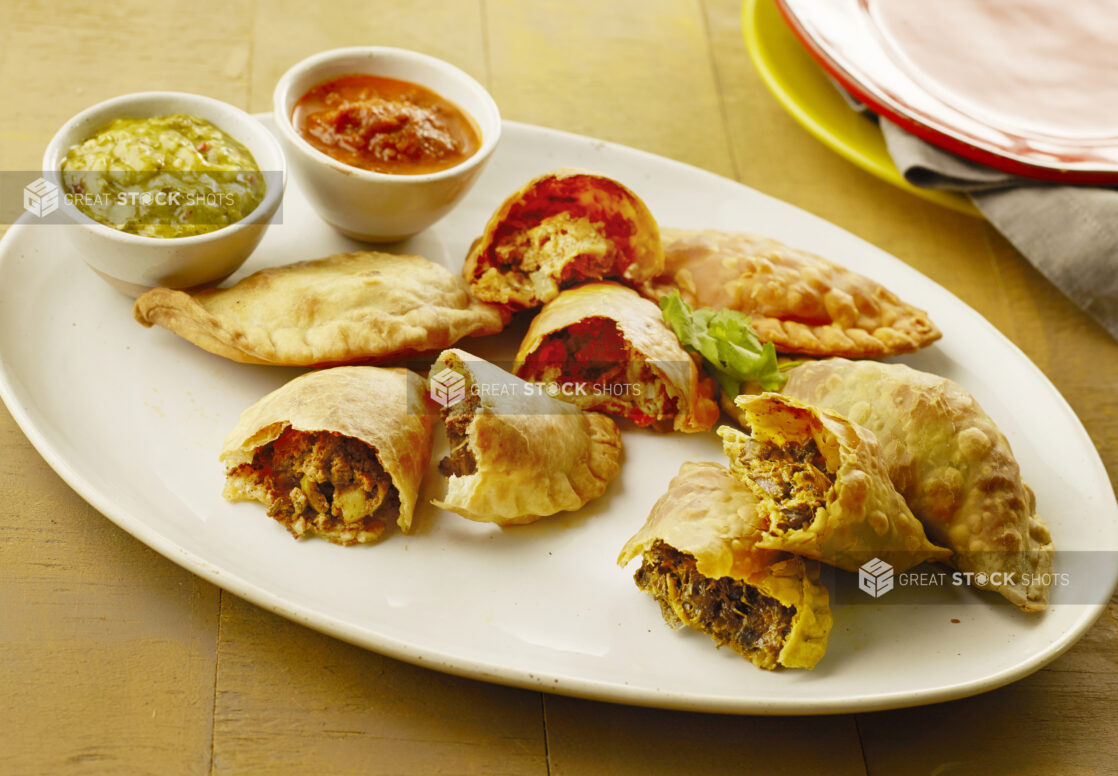 A selection of beef, chicken, and portobello mushroom empanadas with ramekins of red and green salsa on a white oval platter