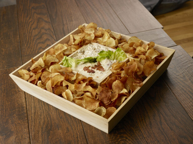 Wooden catering tray of kettle chips with two different dips divided by lettuce on a wooden table