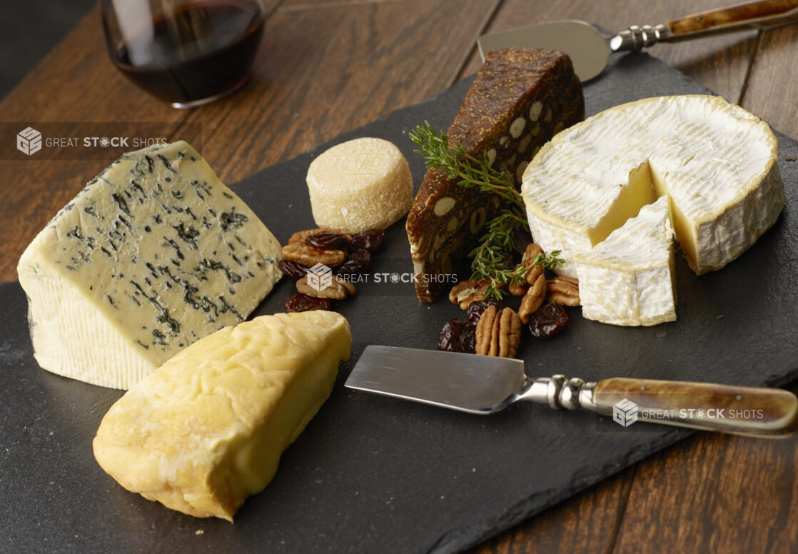 Selection of various cheeses on a slate board with pecans and dried cranberries with a glass of red wine in the background on a wooden table