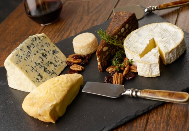 Selection of various cheeses on a slate board with pecans and dried cranberries with a glass of red wine in the background on a wooden table