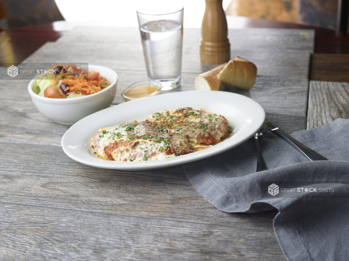 Two chicken parmesan cutlets on a white plate with a side salad and fresh bread in the background on a wooden table