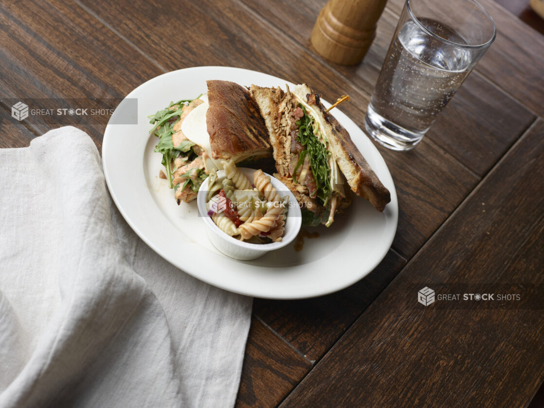 Grilled chicken panini sandwich with arugula, tomatoes, cheese and sauce on white plate with a side pasta salad and glass of water on a wood table