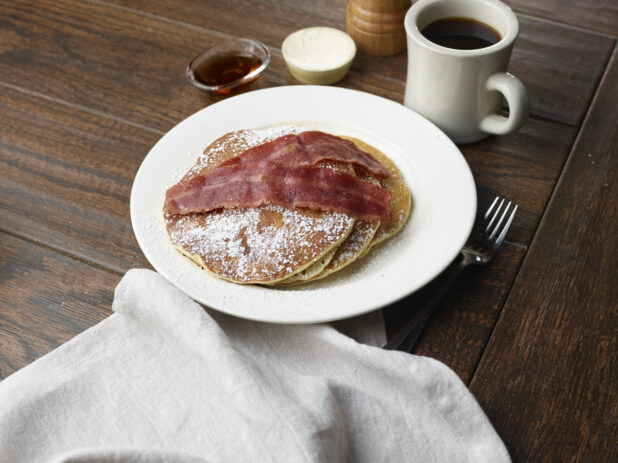 Plate of pancakes topped with icing sugar and bacon with a black coffee, butter and syrup in the background on a wooden table
